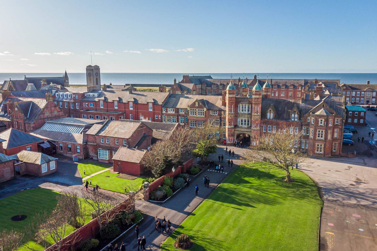 Aerial view of a red-brick UK boarding school campus beside the sea, with lawns and pupils walking between buildings