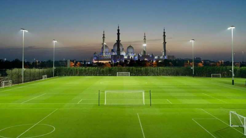Floodlit football pitch at Dubai Sports City at dusk with city skyline in the background